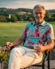 An older man sits in a black metal chair on a stone terrace, holding a cigar and a glass of amber liquor. He is wearing a vibrant turquoise short-sleeve polo shirt with a bold magenta and burgundy tropical floral pattern, paired with beige trousers.