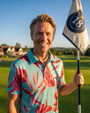 A smiling man on a golf course wearing a light blue polo shirt with a bold red and pink floral pattern, holding a white golf flag with a blue '18' logo.
