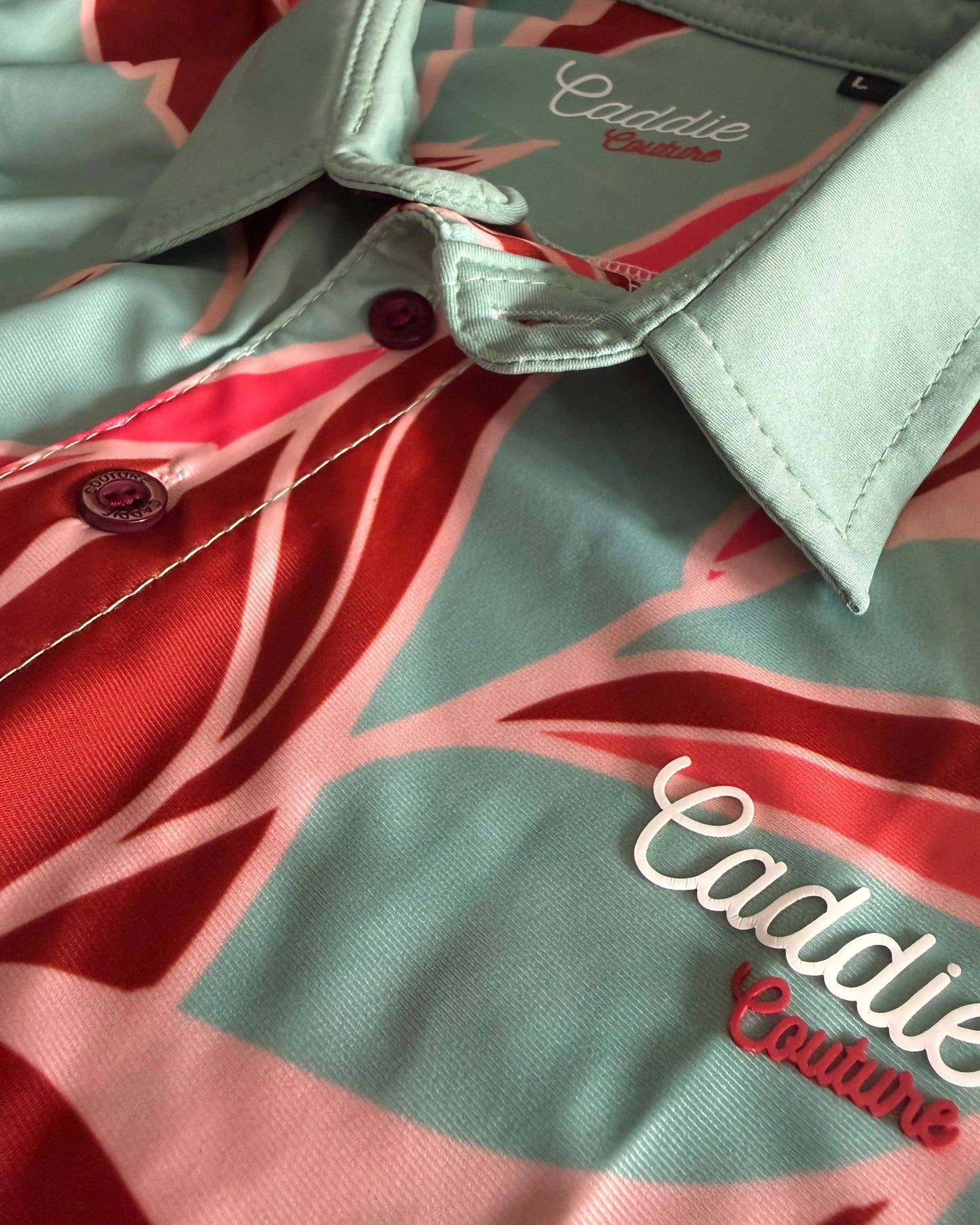 Close-up of a collared button-up shirt with a mint green base and a red and pink tropical leaf pattern. The brand name 'Caddie Couture' is embroidered in white and red on the chest, and the brand name is also visible inside the collar.