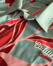 Close-up of a collared button-up shirt with a mint green base and a red and pink tropical leaf pattern. The brand name 'Caddie Couture' is embroidered in white and red on the chest, and the brand name is also visible inside the collar.