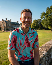 A smiling man wearing a light blue and pink tropical floral patterned polo shirt, standing outdoors on a sunny day next to a stone wall with a large stone mansion and green lawn in the background.