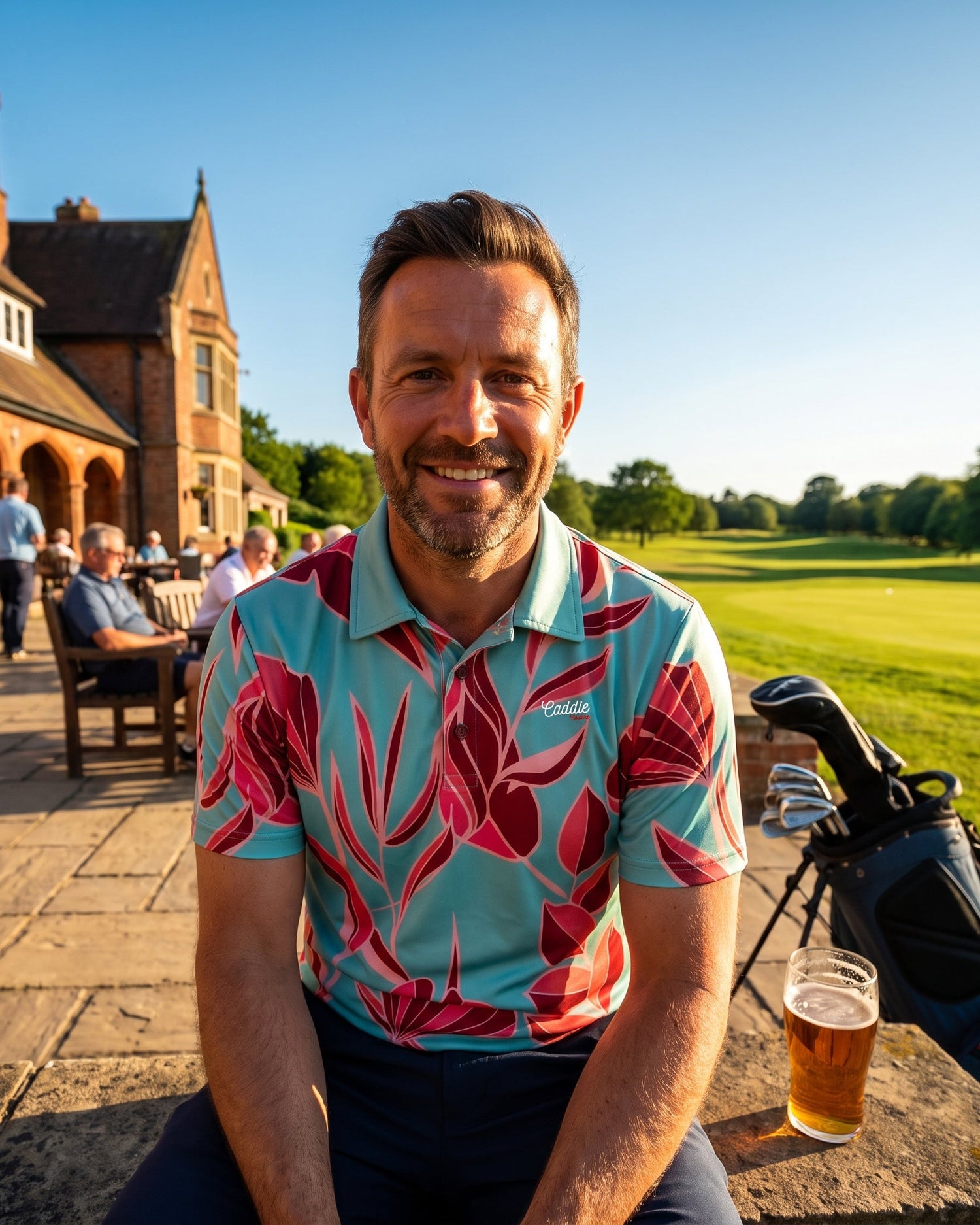A smiling man wearing a light blue polo shirt with a bold red and pink floral pattern sits outdoors on a stone ledge in front of a clubhouse. A glass of beer is on the table next to him, with a golf bag and clubs behind him, and a golf course in the background under a clear blue sky.