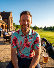 A smiling man wearing a light blue polo shirt with a bold red and pink floral pattern sits outdoors on a stone ledge in front of a clubhouse. A glass of beer is on the table next to him, with a golf bag and clubs behind him, and a golf course in the background under a clear blue sky.