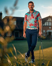 A man wearing a turquoise short-sleeve polo shirt with a red and pink floral pattern, navy trousers, and white golf shoes walks on a golf course holding a club, with a large house in the background.