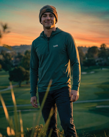 Man standing on a golf course at sunset wearing a green jacket and beanie.