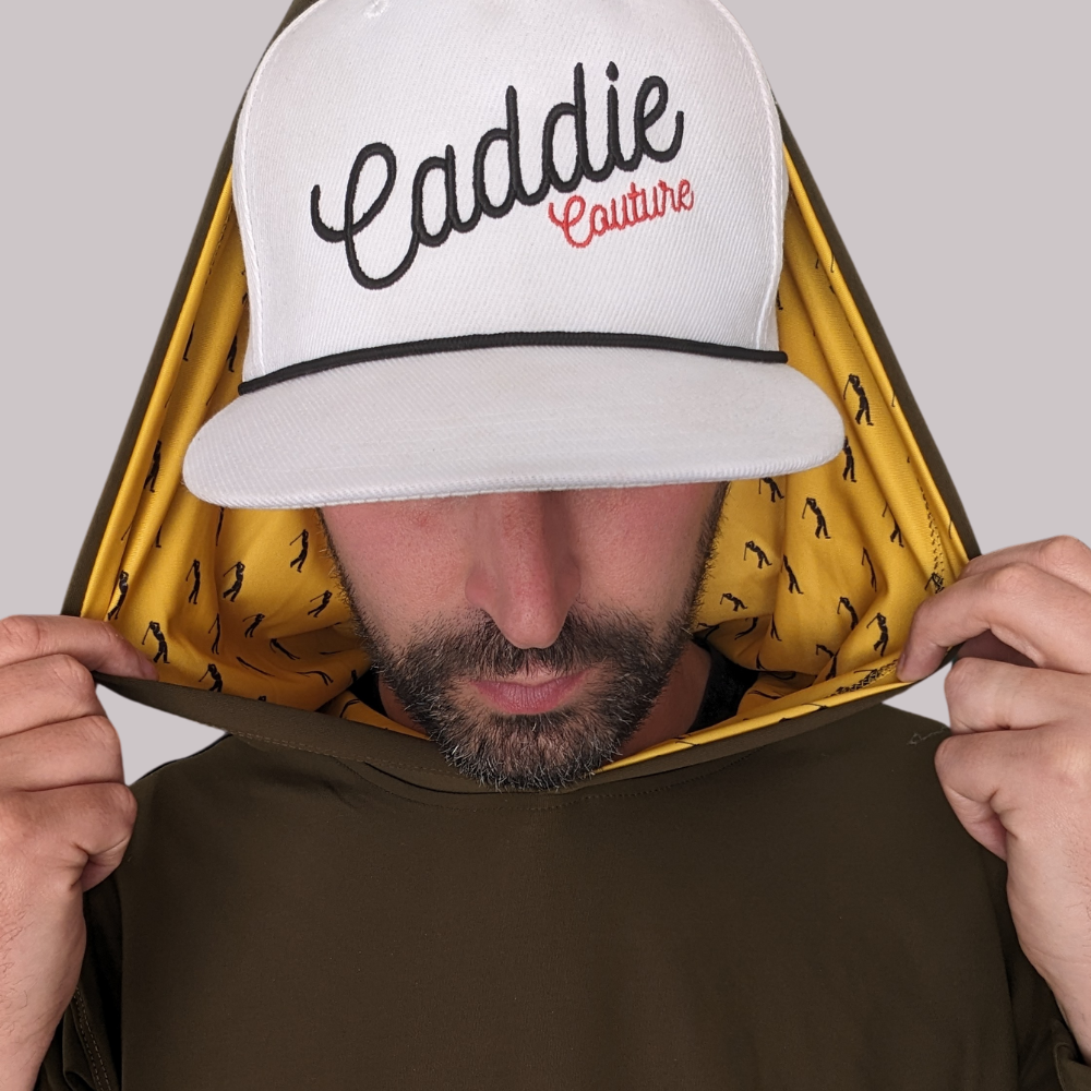 A person wearing a white golf hoodie with 'Caddie Couture' written on the cap in red. The hoodie has an olive green body with yellow golfer design details.