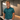 A smiling man wearing a teal and green patterned polo shirt with a dark collar and sleeve cuffs, standing in a modern indoor setting with wood-paneled walls.