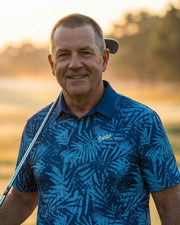 Man in a blue patterned shirt holding a golf club on a golf course at sunset.