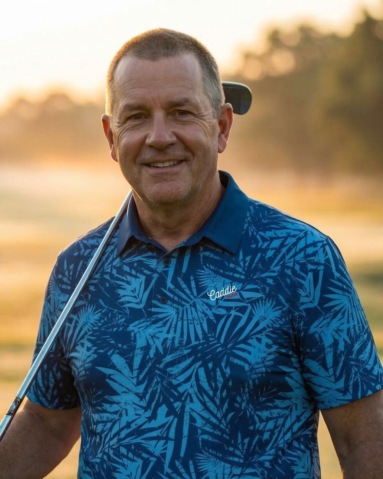 Man in a blue patterned shirt holding a golf club on a golf course at sunset.