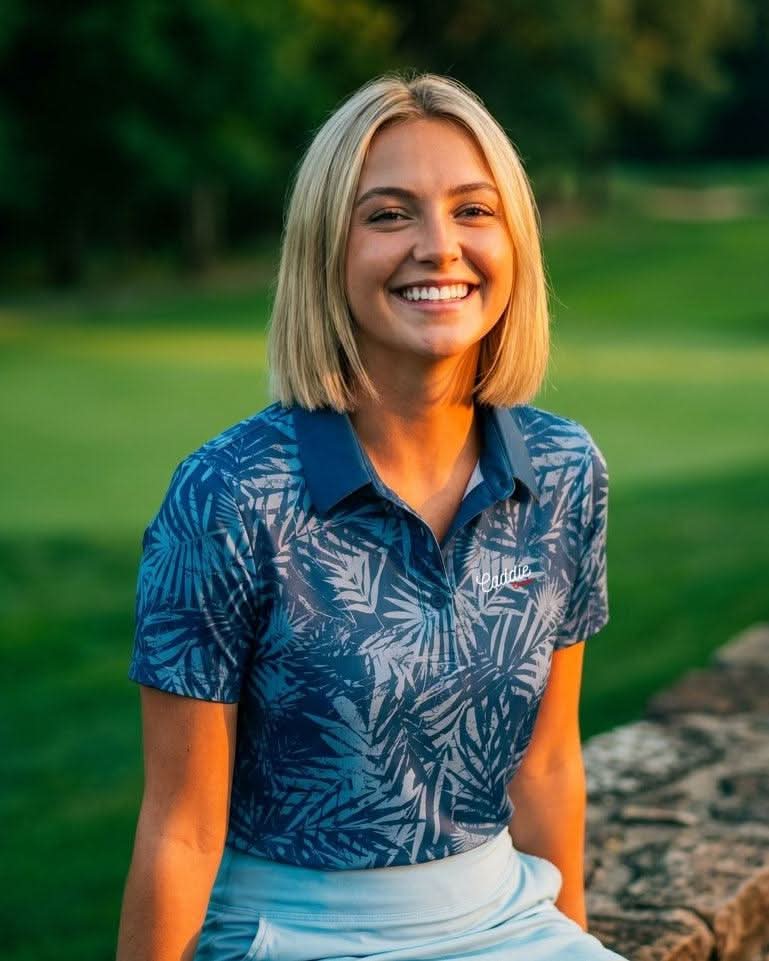 Woman in a blue patterned shirt sitting on a stone wall with a golf course background