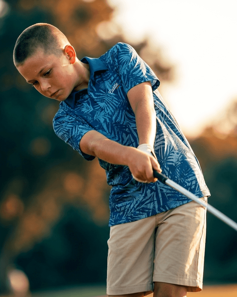 Young boy in a blue patterned shirt and beige shorts holding a golf club on a blurred natural background