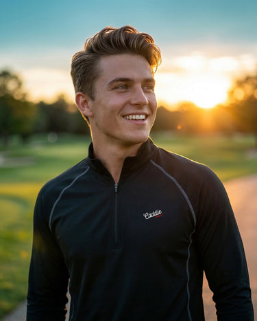 Man wearing a black jacket with a logo, standing outdoors with a sunset in the background