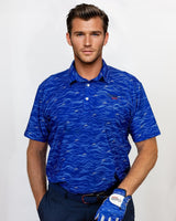 Man wearing a blue patterned golf polo shirt and navy pants, holding a golf club on a white background
