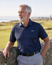 An older man with gray hair and a beard stands outdoors on a golf course, wearing a navy blue polo shirt with a small embroidered logo on the chest, light gray trousers, and leaning against a stone wall.
