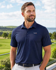 A man wearing a navy blue short-sleeve polo shirt with a small embroidered logo on the chest, standing on a balcony overlooking a golf course.