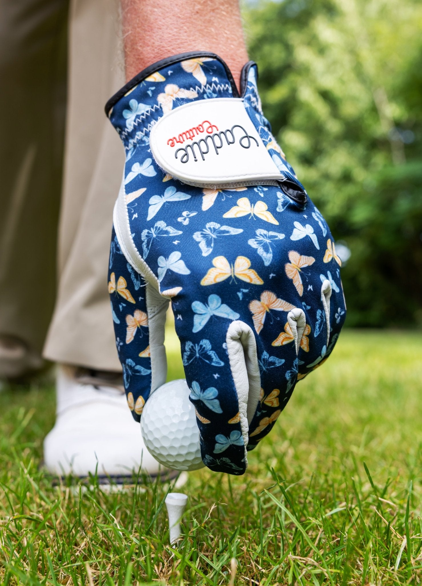 Golf glove with butterfly pattern holding a golf ball on grass