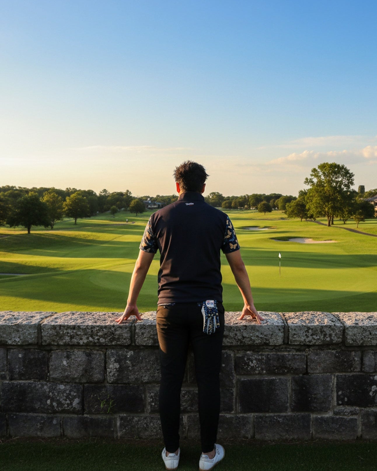 Person standing on a golf course looking over a green with trees and clear sky.
