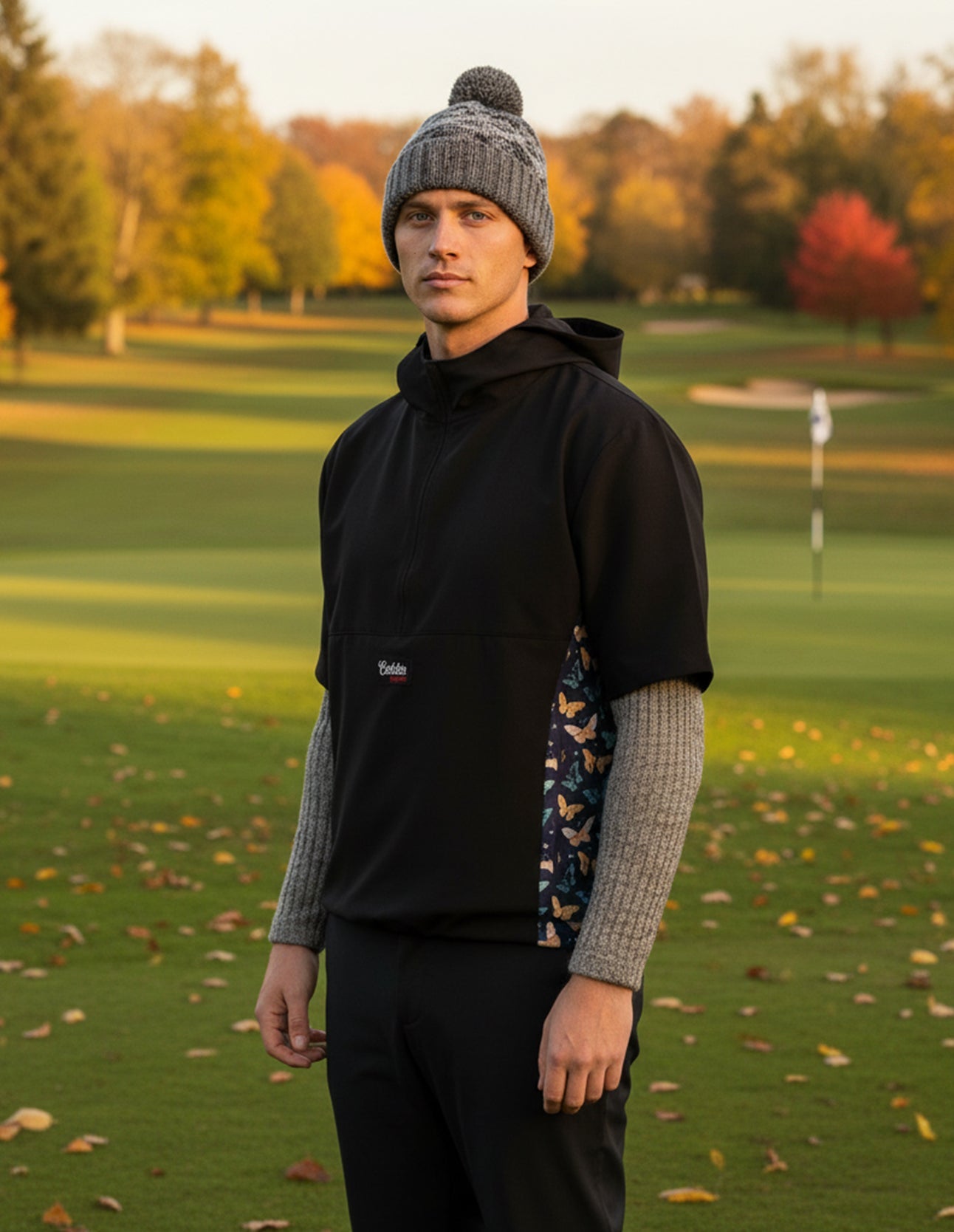 Person standing on a golf course wearing a butterfly windbreaker and gray beanie.
