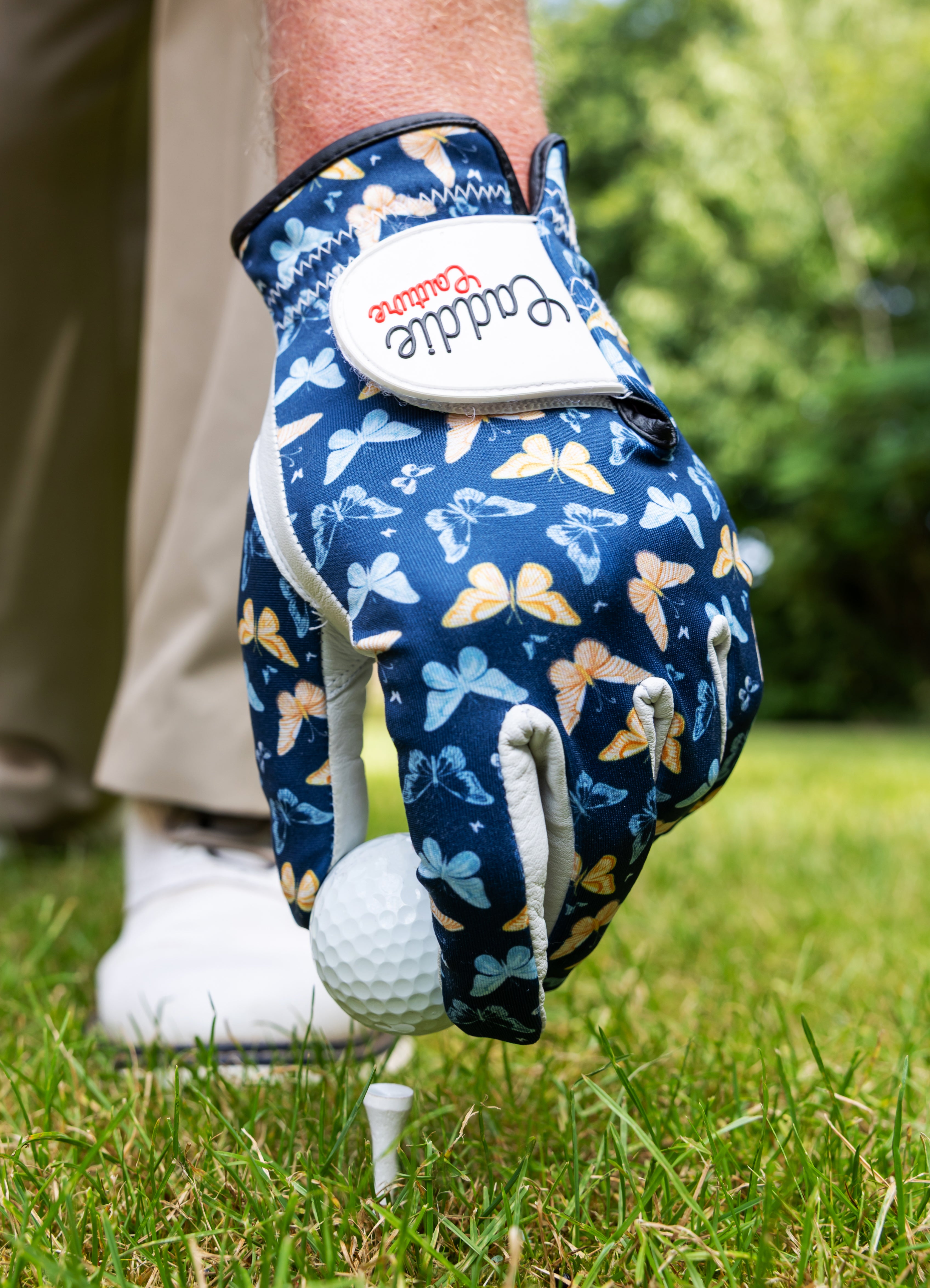 Golf glove with butterfly pattern holding a golf ball on grass