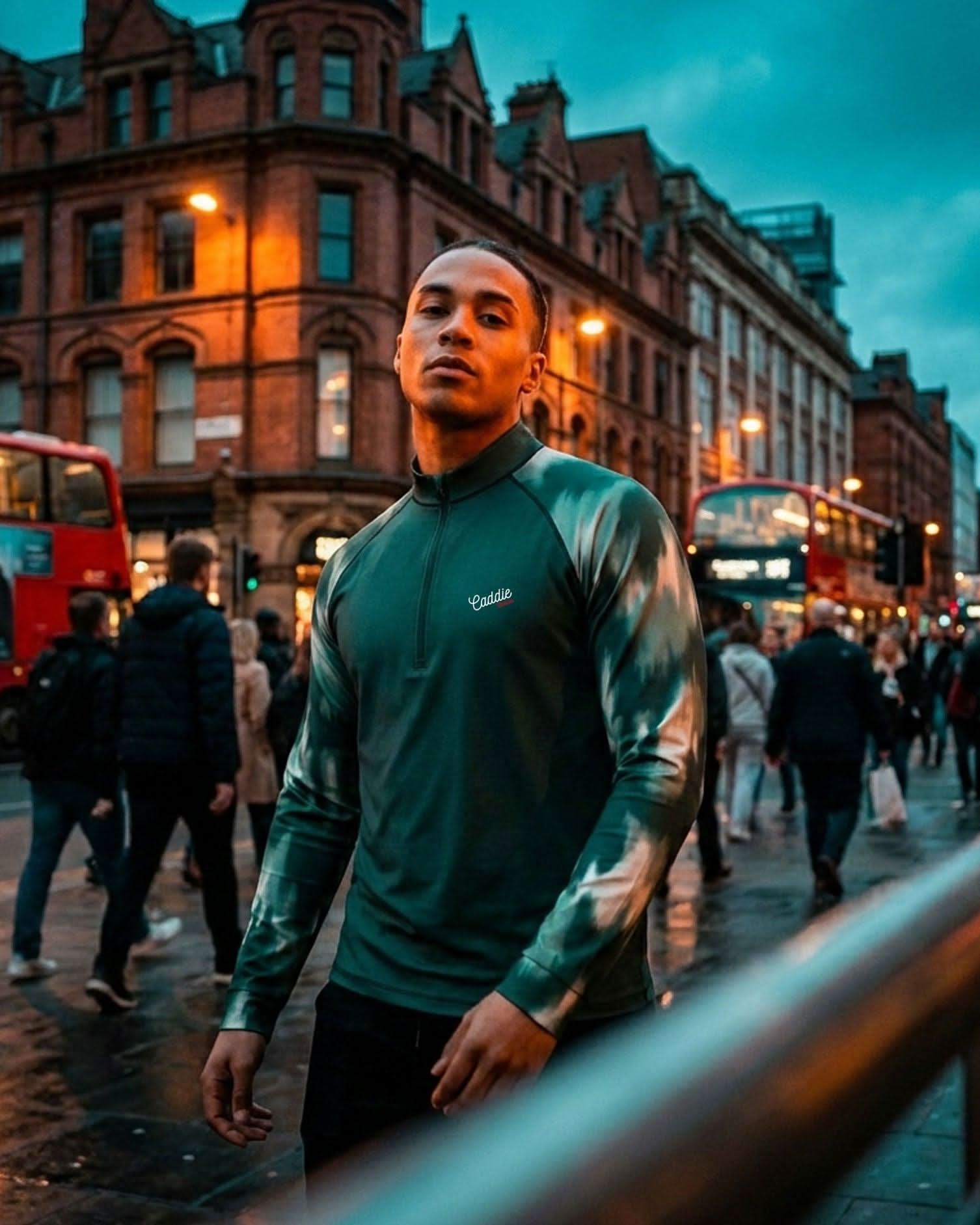 Man in a green jacket standing on a busy city street with red double-decker buses and pedestrians.