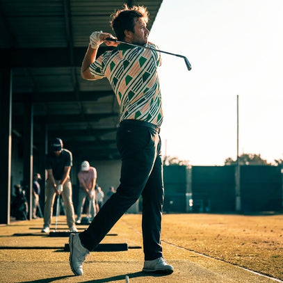 man at a driving range swinging golf club wearing green geo golf polo