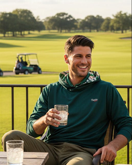 Man sitting on a golf course holding a glass of water with golf carts in the background wearing green geo golf hoodie