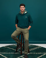 Man sitting on a stool wearing a green geo golf hoodie and khaki pants against a teal wall.