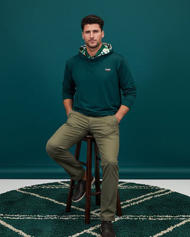 Man sitting on a stool wearing a green geo golf hoodie and khaki pants against a teal wall.