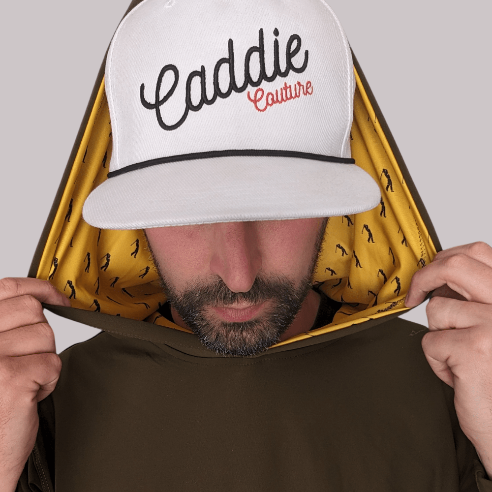 A person wearing a white golf hoodie with 'Caddie Couture' written on the cap in red. The hoodie has an olive green body with yellow golfer design details.
