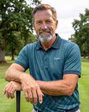 A smiling middle-aged man with a graying beard stands on a golf course, leaning on a golf club. He is wearing a teal polo shirt with a small white embroidered logo on the left chest that reads 'Caddie'.