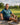 A smiling man sits on a wooden bench at a golf course, wearing a teal polo shirt, navy shorts, and athletic shoes, with a green golf course and trees in the background.