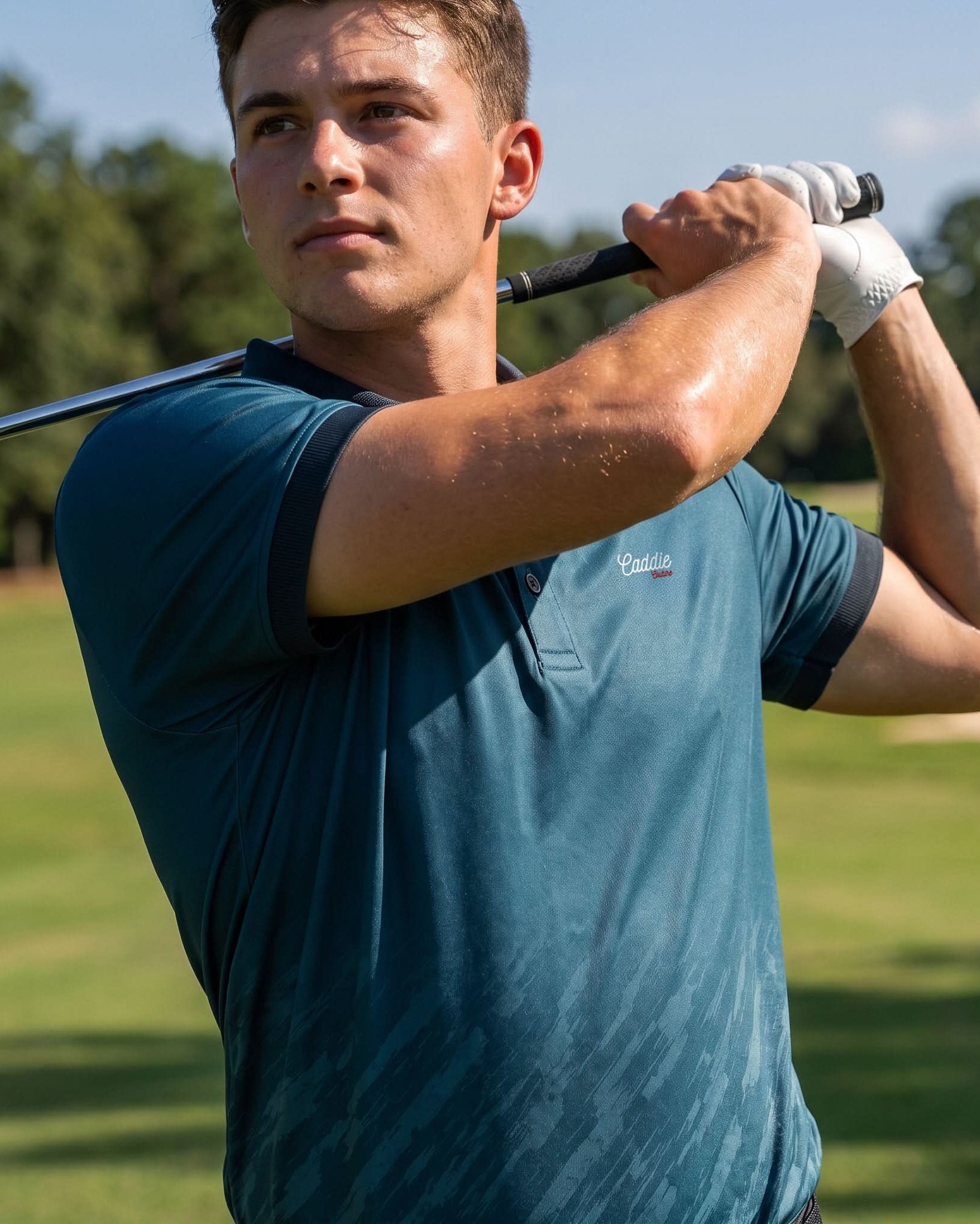 A man on a golf course wearing a teal blue short-sleeve polo shirt with a small white embroidered logo on the chest, holding a golf club in a mid-swing pose.