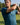 A man on a golf course wearing a teal blue short-sleeve polo shirt with a small white embroidered logo on the chest, holding a golf club in a mid-swing pose.