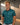 A smiling man wearing a teal and green patterned short-sleeve polo shirt with a dark collar and cuffs, standing in a modern indoor setting with wood-paneled walls and warm lighting.