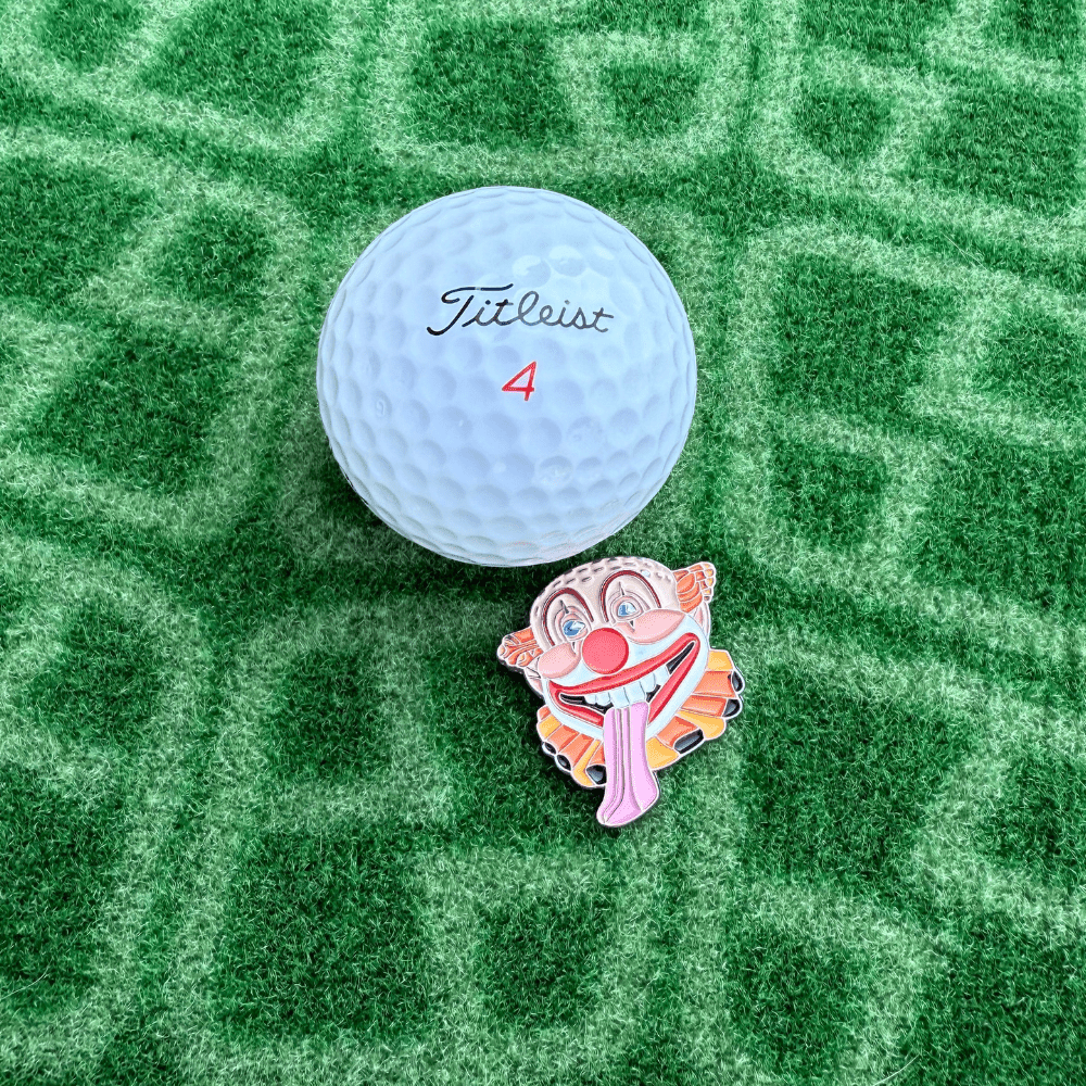 Golf ball with 'Titleist' branding on a green textured surface with Clown Golf Ball Marker Next to it