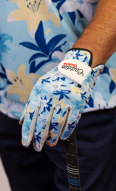 Golf glove with floral pattern worn by a person, holding a golf club, against a blue floral background.