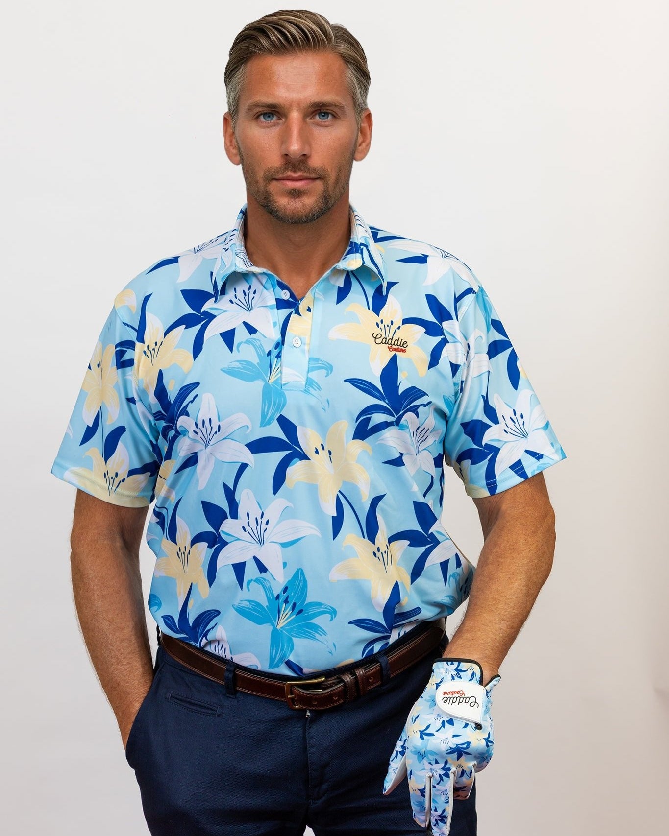 Man wearing a blue floral golf shirt and dark pants, holding a golf club with a white background