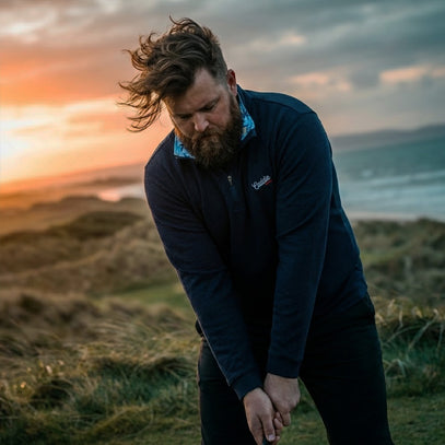 Man in a dark jacket standing on a grassy hill with a sunset or sunrise in the background.