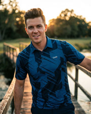 Man wearing a blue patterned shirt standing on a wooden bridge by a body of water.