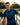 Man wearing a blue patterned shirt standing on a wooden bridge by a body of water.