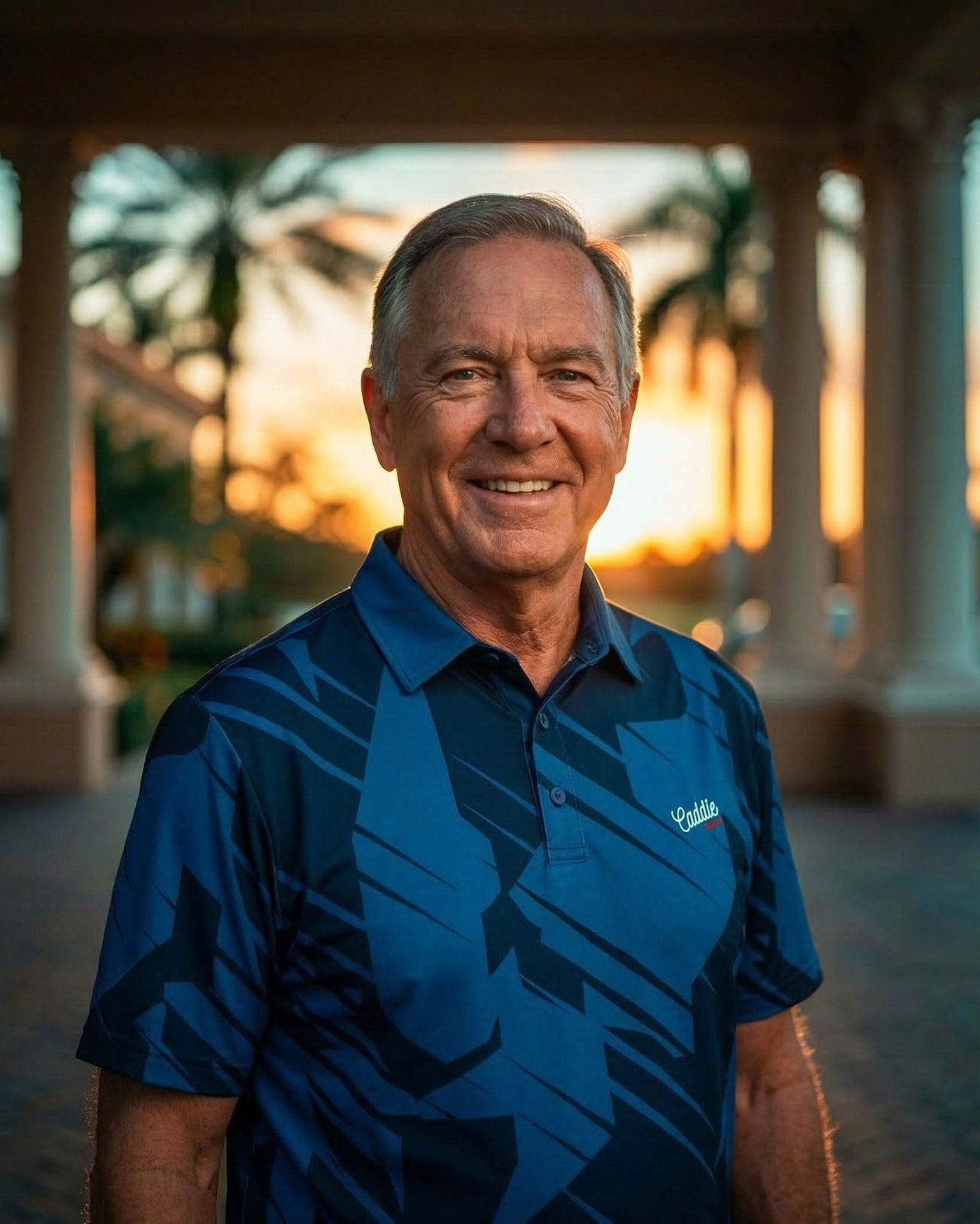 Man wearing a blue polo shirt with a logo, standing in front of a blurred outdoor setting with columns and greenery.