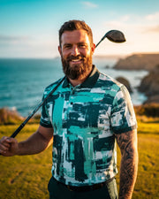 A smiling bearded man wearing a teal, white, and black abstract-patterned polo shirt stands on a grassy golf course holding a golf club, with the ocean and cliffs in the background.