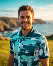 A smiling man wearing a teal, green, and white abstract print polo shirt, standing outdoors on a grassy hill with a coastal ocean and cliffs in the background.