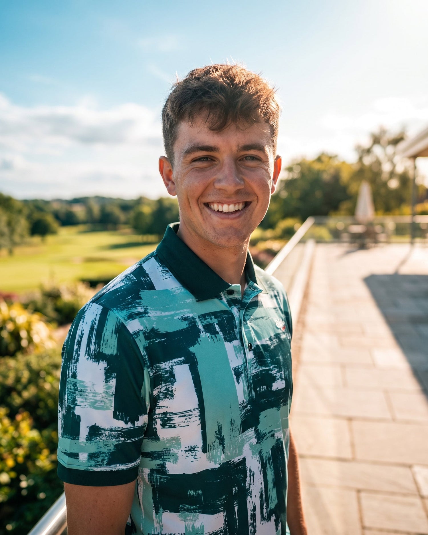 A smiling man wearing a green and white abstract-patterned polo shirt, standing outdoors on a sunny day with a golf course and patio visible in the background.