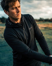 Man wearing a black jacket with a brand logo, standing in a field with a cloudy sky.