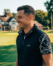 Man in a black and white patterned shirt standing on a golf course with houses in the background