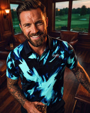 Man wearing a blue patterned shirt with a logo, holding a drink indoors with a golf course view.