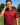 A man on a golf course wearing a red and black patterned polo shirt with a visible 'Caddie Couture logo, holding a golf laser sight to his eye.