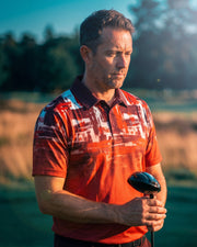 A man on a golf course wearing a red and orange patterned polo shirt, holding a golf club.