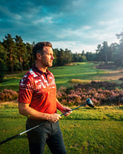 A man wearing a red and white patterned polo shirt and dark trousers stands on a golf course holding a golf club, with trees and a blue sky in the background.